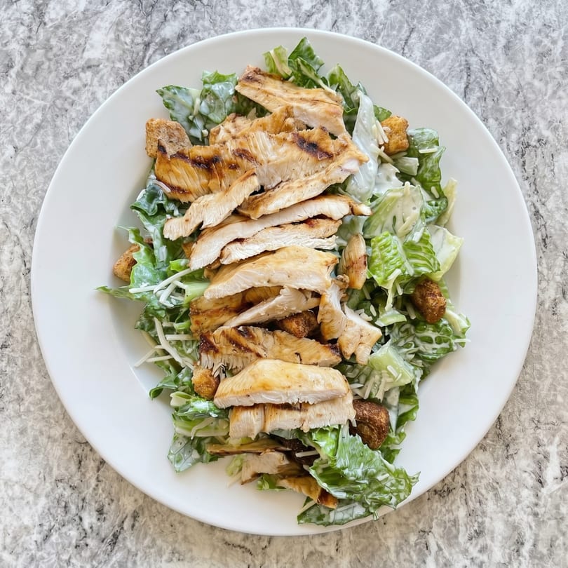Grilled chicken Caesar salad with romaine lettuce, croutons, and dressing on a white plate.